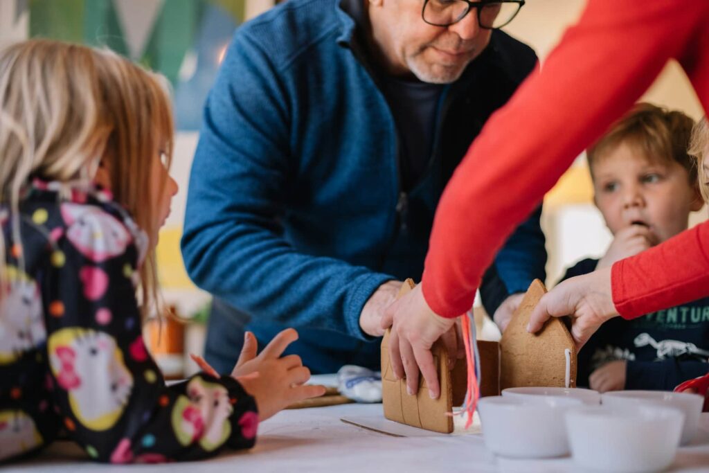 Two people making gingerbread house and three children watching them