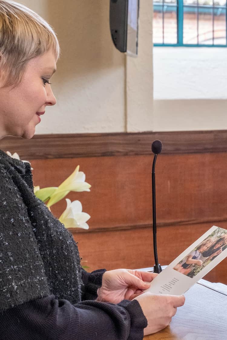 A woman holding an order of service stands at a lectern at a funeral ceremony